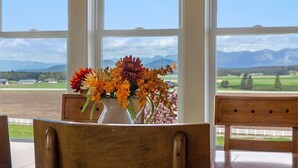 A room with a large window offering a view of mountains and a field. A wooden chair is positioned in front of the window, and a vase with flowers is on the windowsill. - Majestic Mountain Views! (Kalispell)
