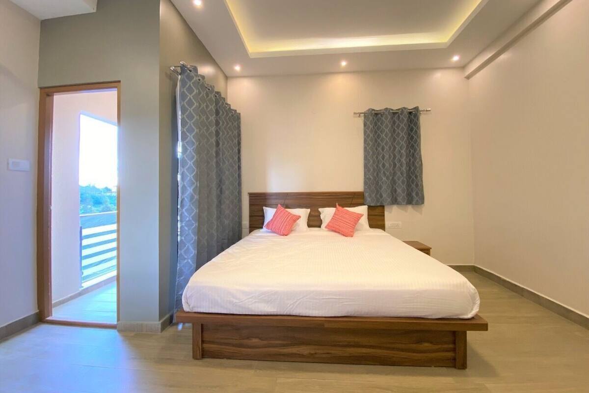 Un dormitorio con cama de madera, dos almohadas, una mesita de noche, un ventanal con cortinas y una puerta que da a un balcón.