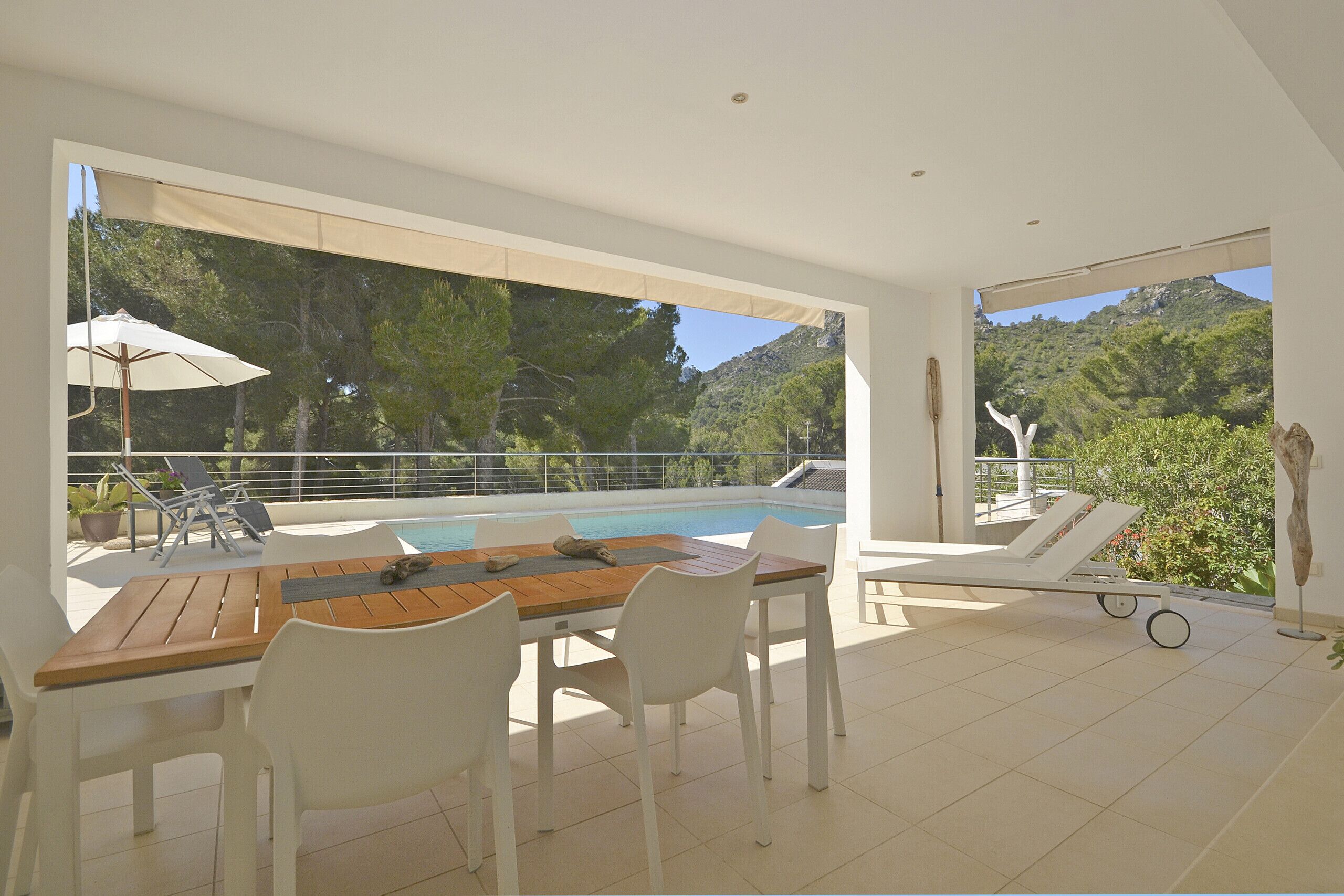 A modern outdoor pool area with a wooden table and white chairs, surrounded by a glass railing and overlooking a mountainous landscape.