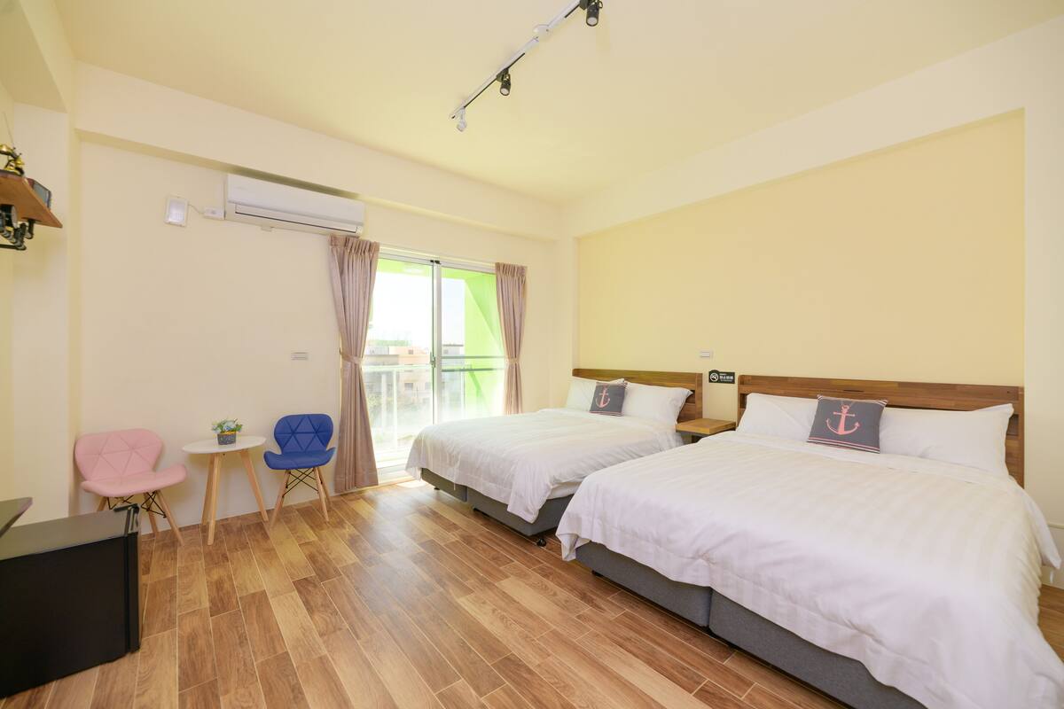 A hotel room with two beds, a small table with chairs, an air conditioning unit, and a view through a glass door.