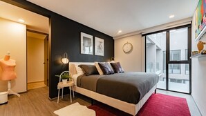 Ein modernes Schlafzimmer mit einem großen Bett, einem roten Teppich, einer weißen Kommode, einer Mannequin und einem großen Fenster mit Blick auf Gebäude.