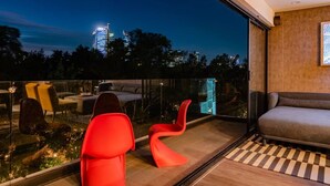 Ein moderner Balkon mit Glasboden, roten Stühlen und einer Couch, mit Blick auf eine beleuchtete Stadtlandschaft bei Nacht.