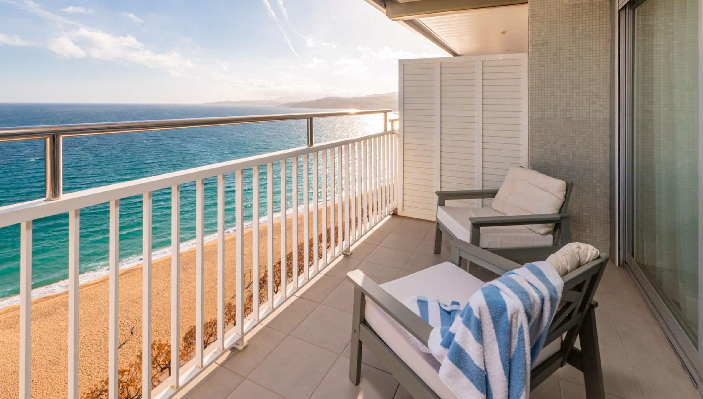 A balcony with a view of the ocean, a chair, a small table, and a tiled floor.