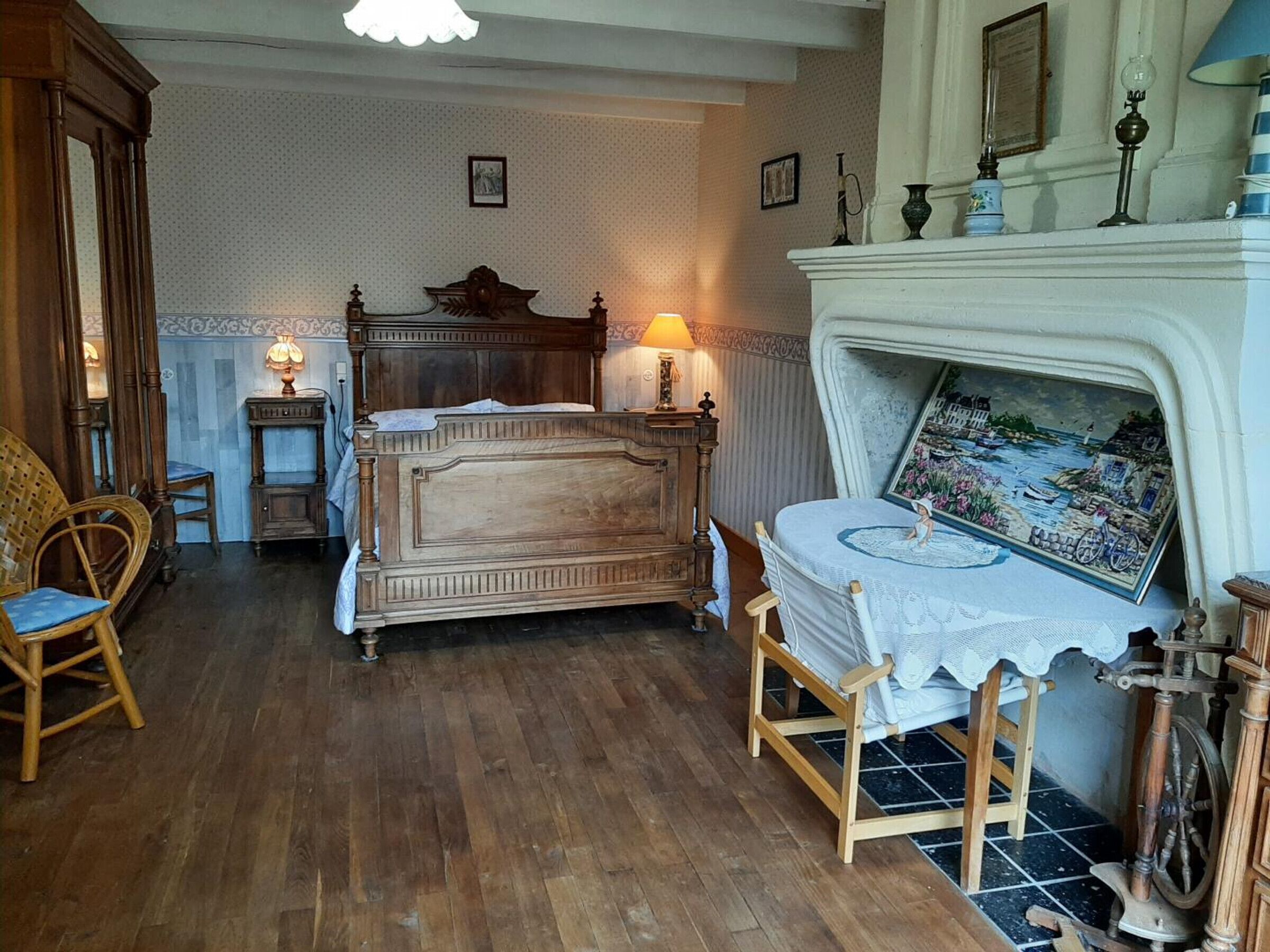 Een klassieke slaapkamer met een houten bed, een klein tafeltje met een lamp, een stoel en een open haard met een schilderij erboven.