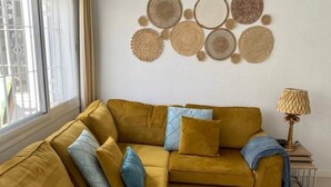 En sofa i senegul farve med blå og grå puder, dekoration på væggen i vævet materiale, et lille bord med en lampe, og et vindue med persienner.