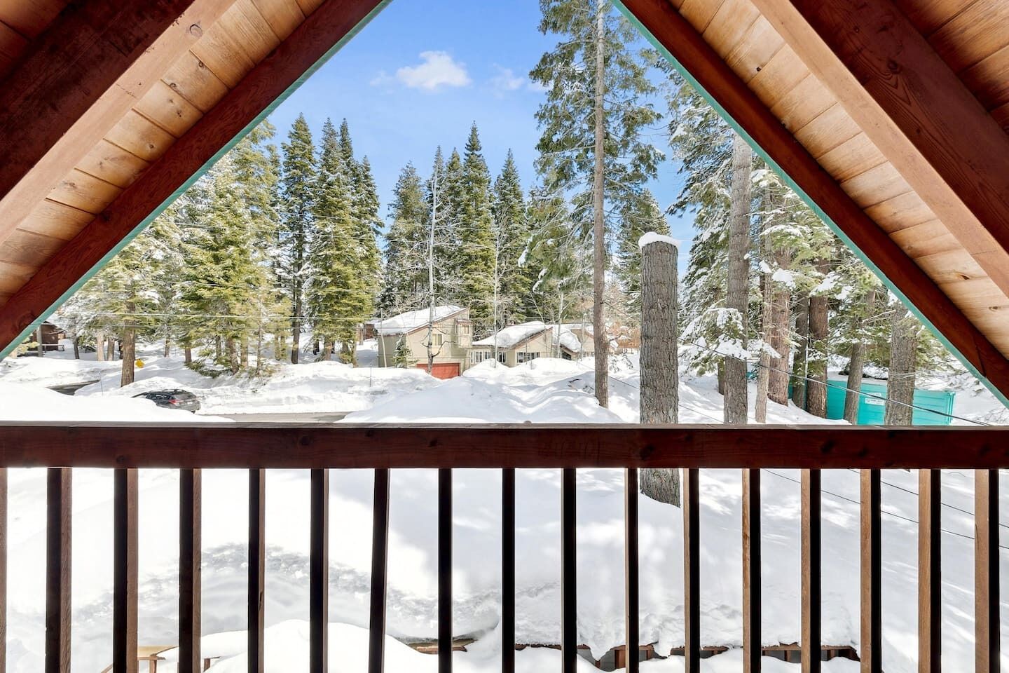 A view from a wooden balcony with a snow-covered landscape, tall trees, and a house in the background.