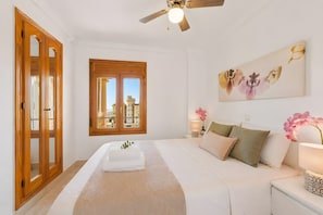 A bedroom with a bed, a window with a view, a ceiling fan, and a painting on the wall.