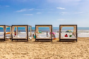 Trois cabanes de plage équipées de lits, une plage de sable et l’océan en arrière-plan.