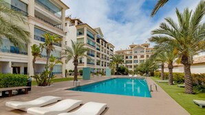 A hotel with a swimming pool, lounge chairs, palm trees, and a well-maintained lawn.