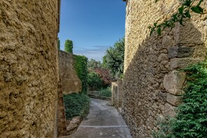 A narrow stone pathway flanked by stone walls, leading to a lush green area with trees and shrubs.