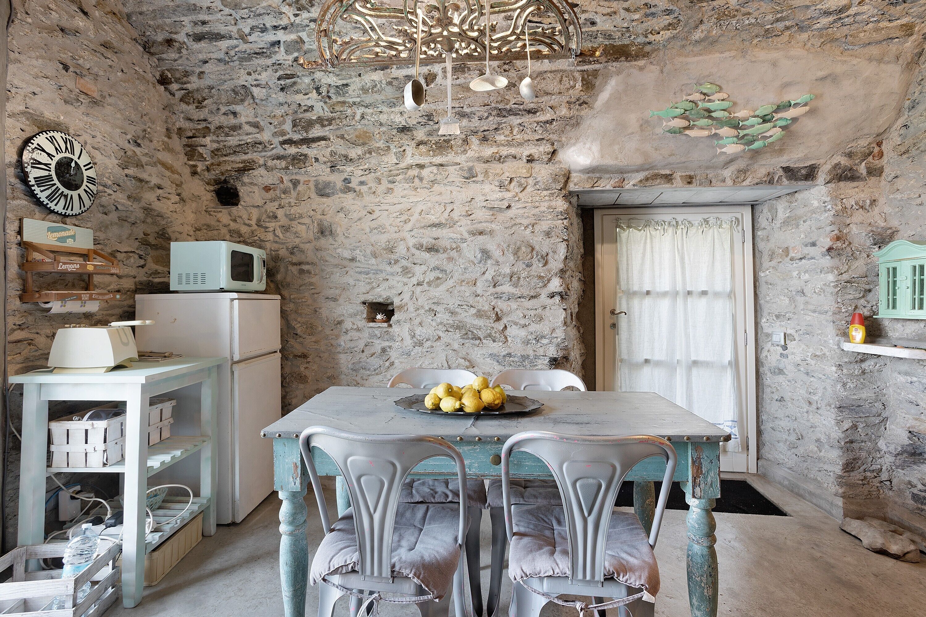 Une pièce rustique aux murs de pierre, comprenant une table à manger, des chaises, un lustre et une horloge.