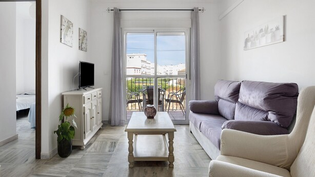 Una sala de estar con sofá, televisor en un mueble, mesa y balcón con mobiliario exterior.