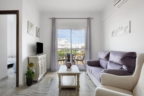 Ein Wohnzimmer mit einer Couch, einem Fernseher auf einem Schrank, einem Tisch und einem Balkon mit Gartenmöbeln.