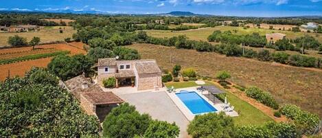 A stone house with a swimming pool, surrounded by greenery and fields.