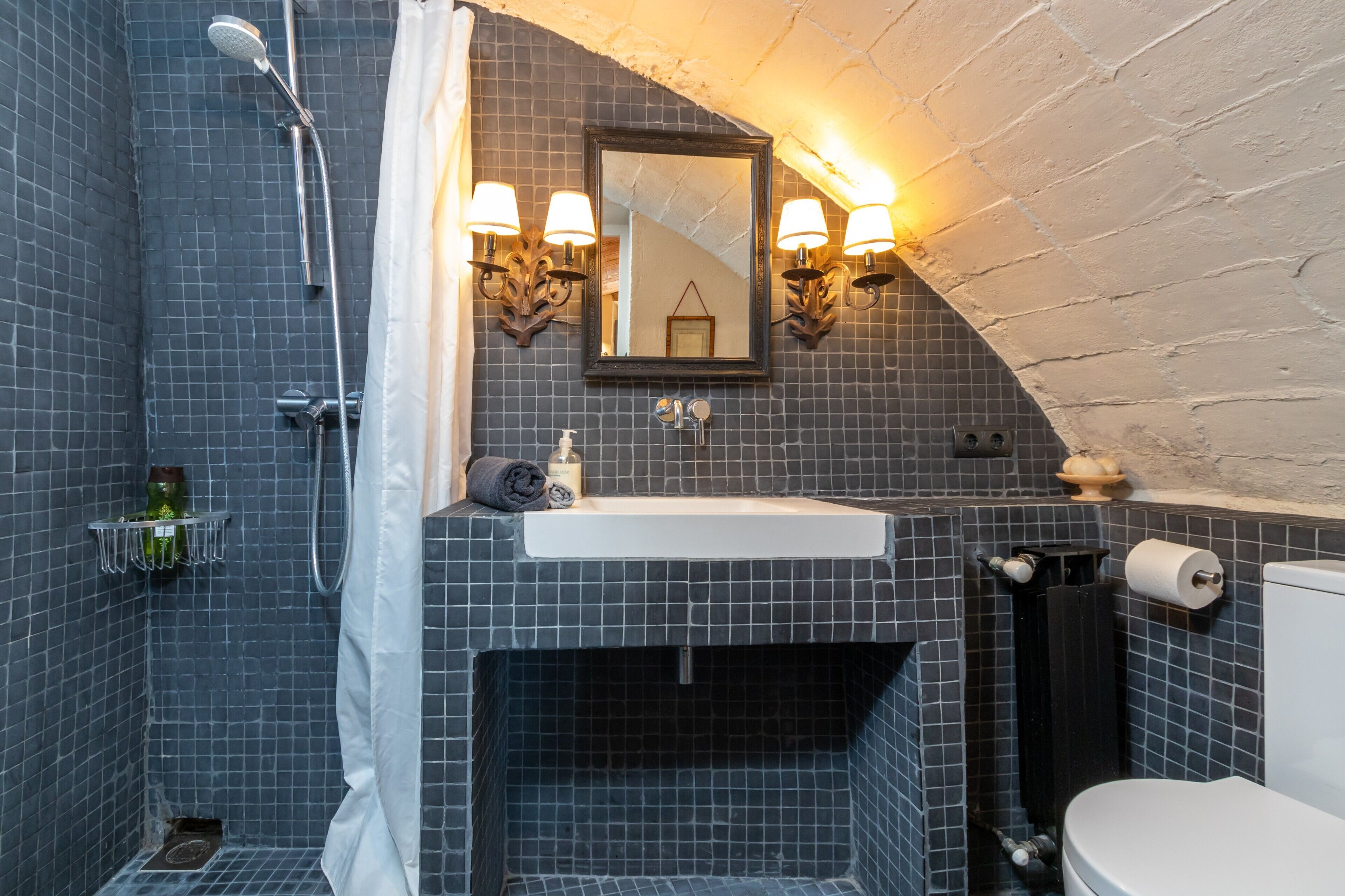 Un baño con ducha, lavamanos, inodoro y espejo con apliques de pared.