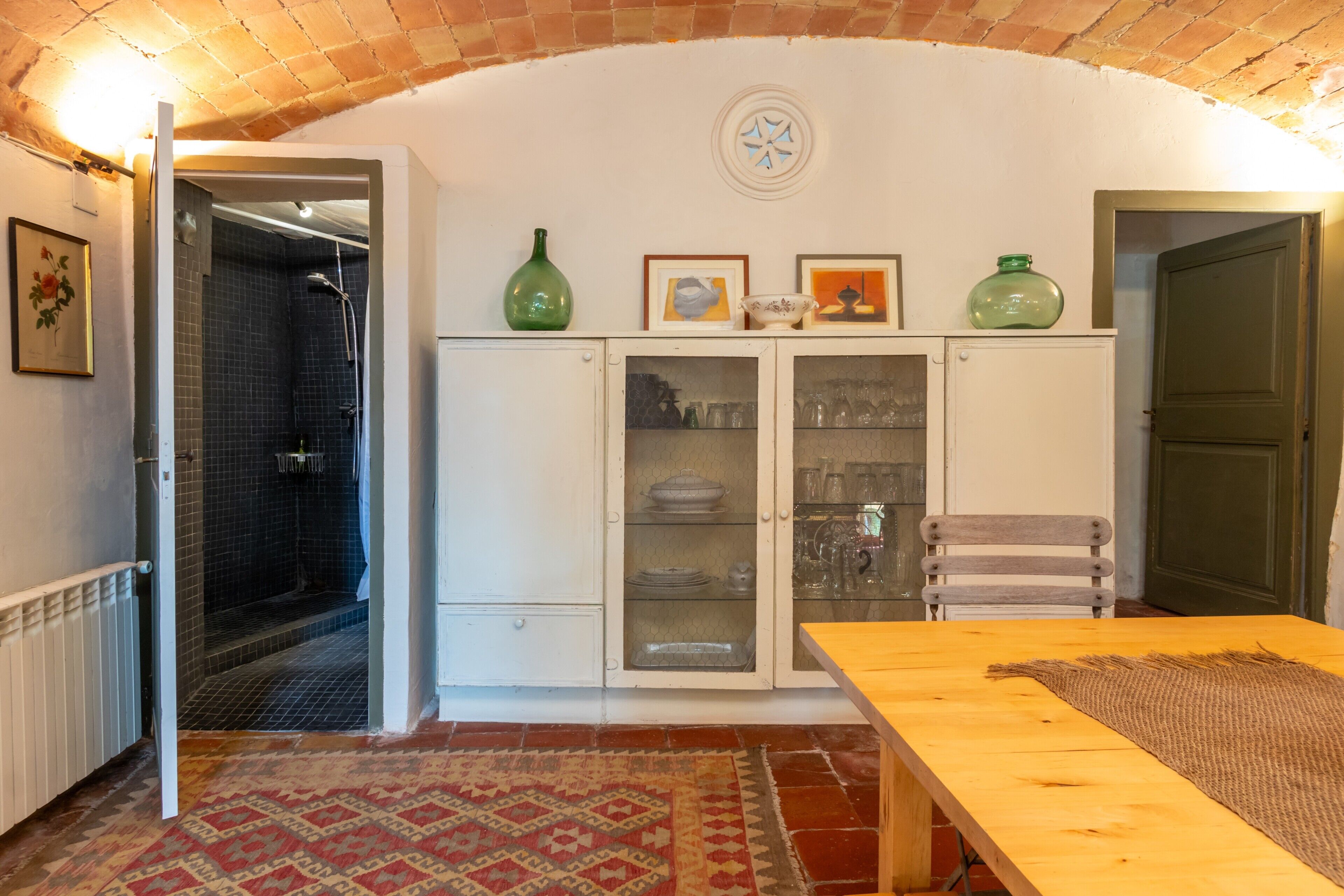 An open doorway leading to a shower, a wooden table with a woven placemat, and a cabinet with glass doors containing dishes and glasses.