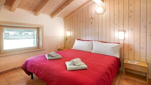 A bedroom with a wooden ceiling, a red bedspread, two white pillows, a wooden nightstand, and a window with lace curtains. - Apartment 'Appartamenti Lorenza 3 Baite Giotto' with Mountain View, Shared Garden and Wi-Fi (Livigno)
