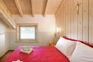 A room with a bed, a window, wooden beams, and a wall-mounted cross.