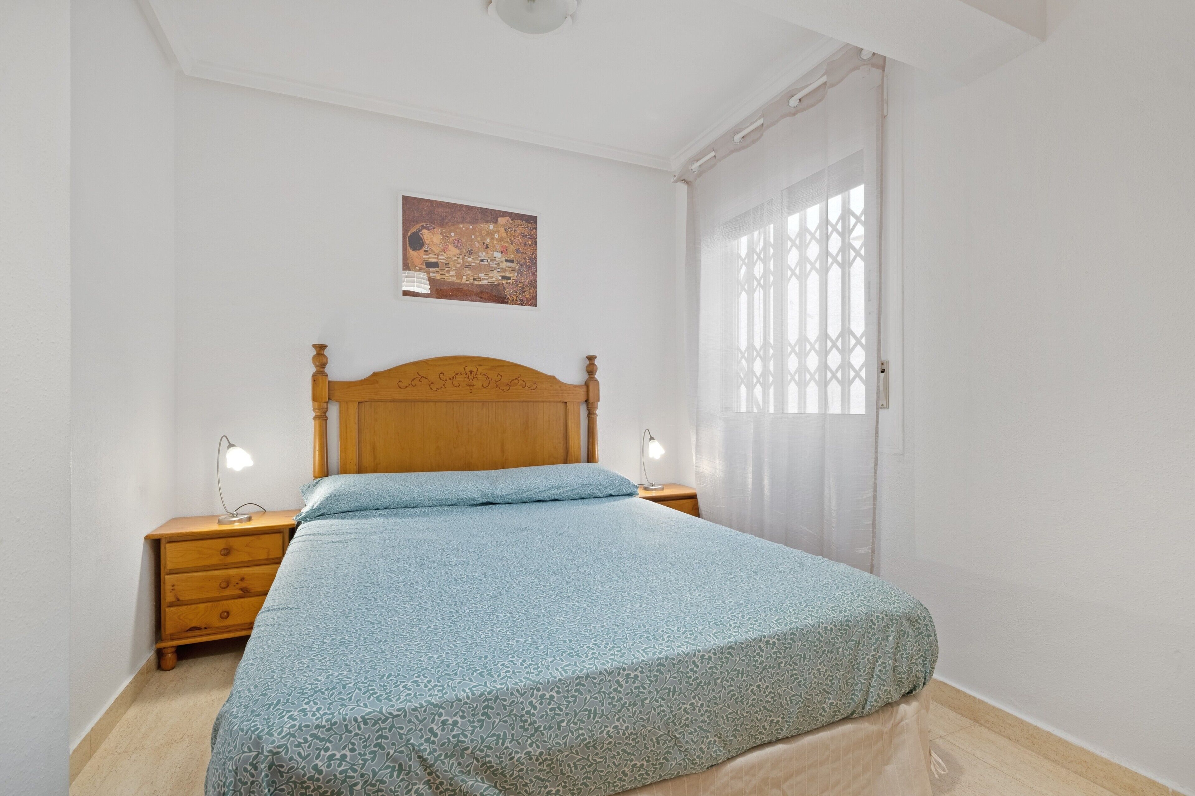 Ein Schlafzimmer mit einem hölzernen Bett, zwei Nachttischen mit Lampen, einem kleinen Gemälde an der Wand und einem Fenster mit weißem Vorhang.