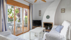 Une chambre avec un lit, une table blanche, une télévision, une cheminée et un balcon agrémenté de chaises et de plantes.