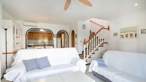 Un salon avec un canapé blanc, une table basse blanche, un ventilateur de plafond et un escalier doté de rampes en bois.