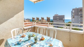 Un balcon avec une table dressée pour deux personnes, des plantes en pot et une vue sur la mer et les bâtiments.