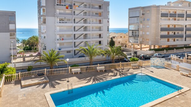 Une piscine avec vue sur la mer, entourée de palmiers et bordée d’un bâtiment blanc doté de balcons.