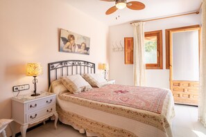 A bedroom with a bed, two bedside tables with lamps, a ceiling fan, a window with curtains, and a wooden door.