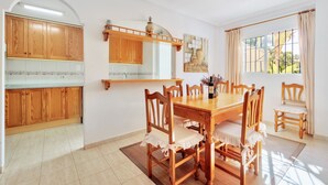 A dining area with a wooden table and chairs, a kitchen with wooden cabinets, a hanging light fixture, and a window with curtains.