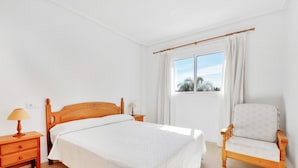 A bedroom with a wooden bed, a nightstand with a lamp, a chair, and a window with curtains.