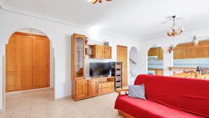 A spacious living area with a red sofa, wooden cabinets, a television, and a kitchen area with wooden cabinets and a hanging light fixture.