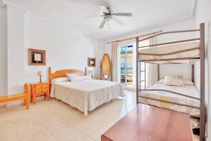 Une chambre avec un lit simple, une table de chevet en bois, un banc, un ventilateur de plafond et un lit superposé.
