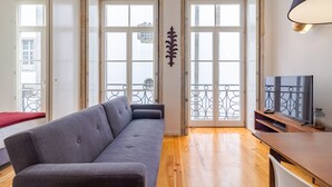 A modern living room with a grey sofa, wooden flooring, a TV stand, and large windows with a balcony view. - Twin Studio with Balcony view Clérigos Tower (Oporto)