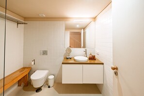 Een moderne badkamer met toilet, wastafel en badkamermeubel, met witte tegels en een houten bankje.