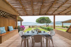 A covered patio with a table set for dining, a seating area with cushions, and a view of the sea and hills.