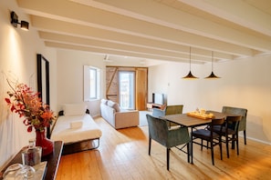 A modern hotel room with a wooden floor, a dining area with a table and chairs, a sofa, a TV, and a large window.