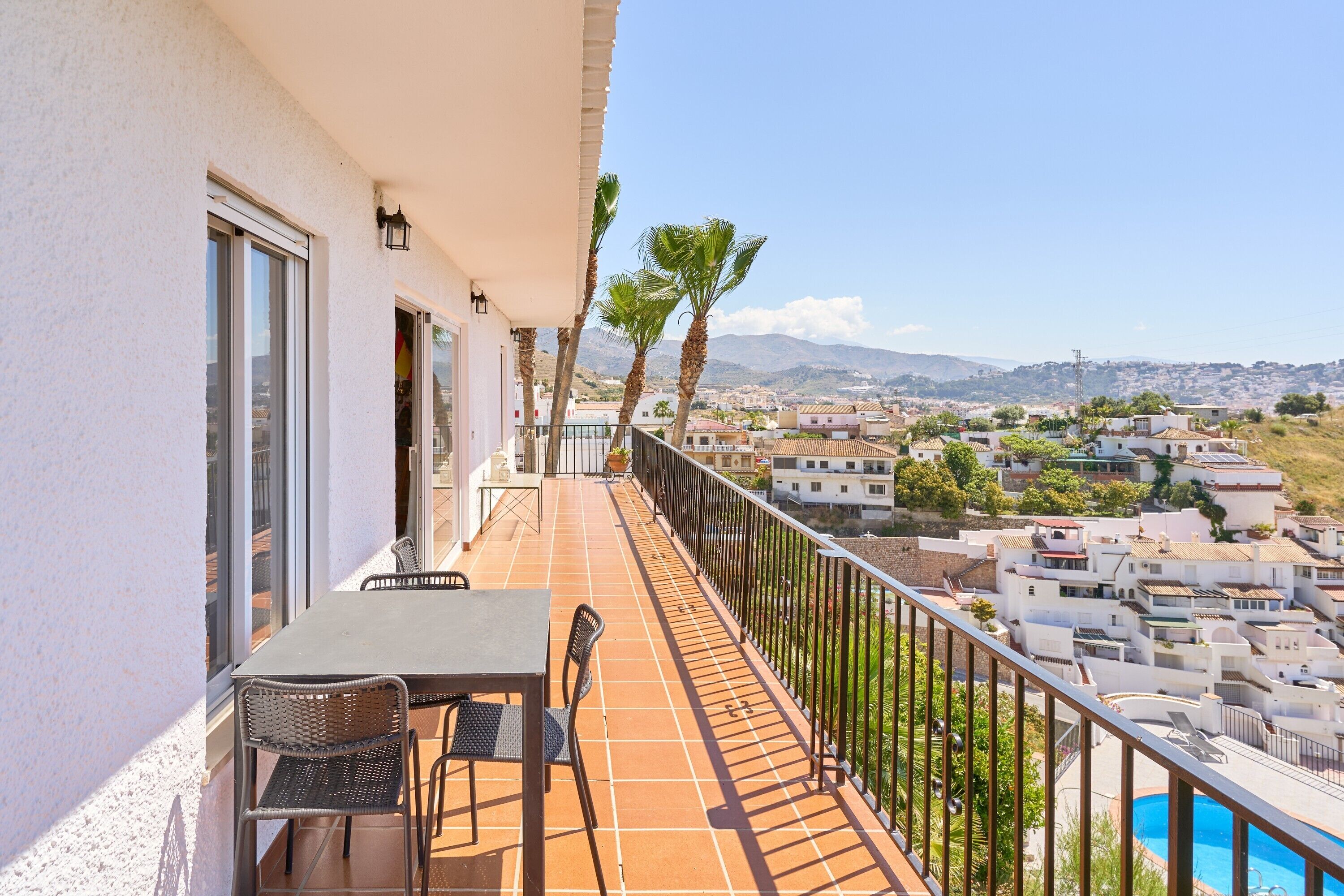 A balcony with a table and chairs, overlooking a residential area with houses and a mountain in the distance.