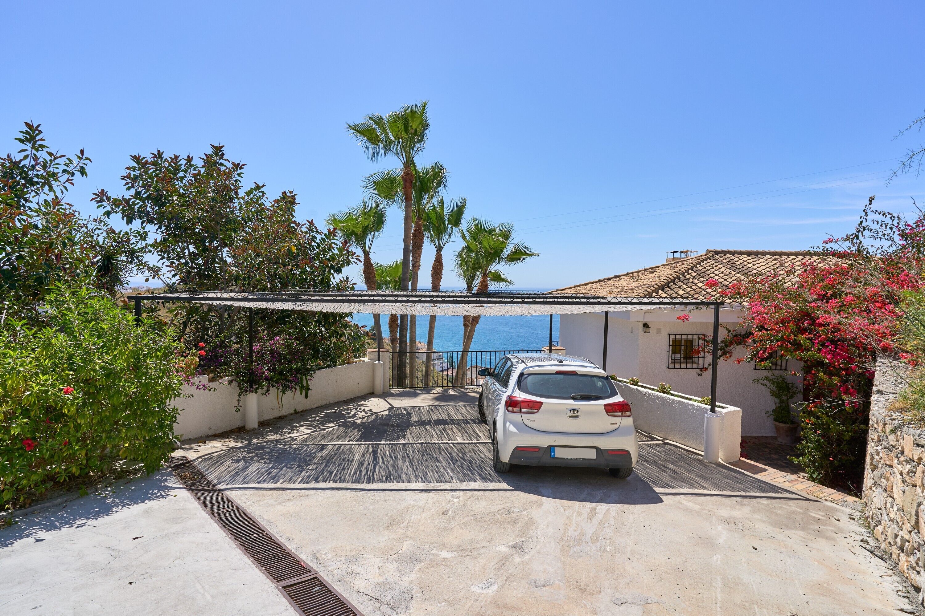 A white car parked in a covered driveway with a view of the sea and palm trees.
