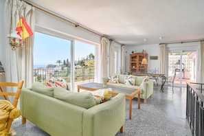 A spacious living area with a sofa, coffee table, and dining table. Large windows offer a view of the outdoors.