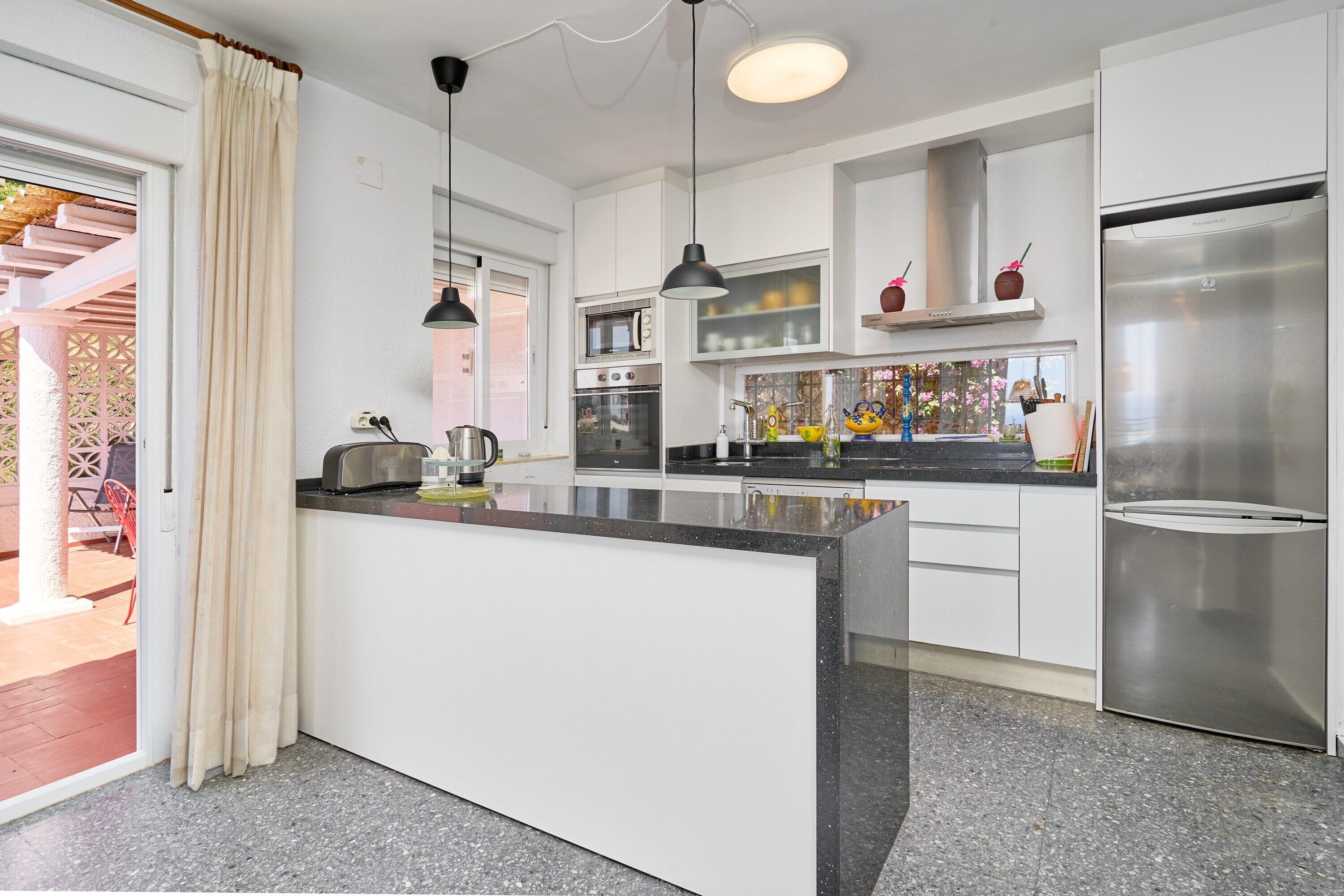 Une cuisine moderne avec des meubles blancs, un îlot central, des appareils en acier inoxydable, des luminaires suspendus et une vue sur une terrasse extérieure.