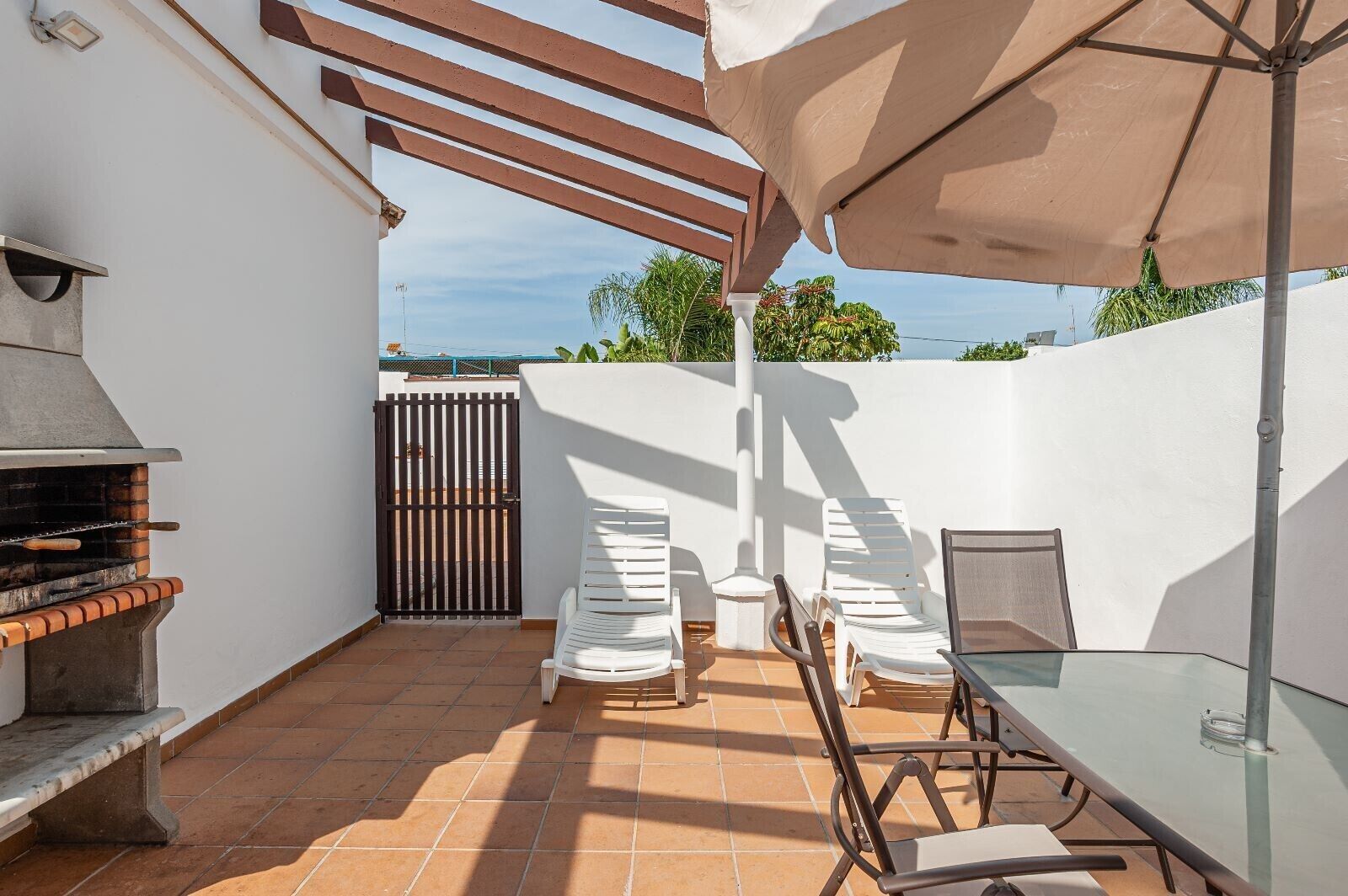 Um pátio externo com churrasqueira, cadeiras brancas, mesa de vidro e uma pérgula com guarda-sol.