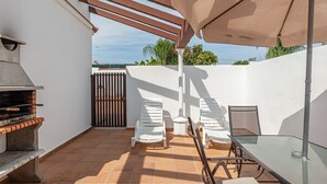 Une terrasse extérieure avec un barbecue, des chaises blanches, une table en verre et une pergola surmontée d’un parasol.