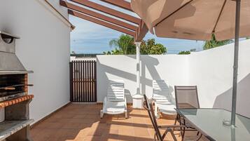 Um pátio externo com churrasqueira, cadeiras brancas, mesa de vidro e uma pérgula com guarda-sol.