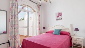 Une chambre à coucher avec un lit, des rideaux, une table de chevet, une lampe, un tableau accroché au mur et une fenêtre donnant sur un paysage.