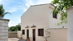 Ein Gebäude mit flachem Dach, zwei Türen und Fenstern mit Rollläden. Im Hintergrund sind Kübelpflanzen und eine Steinmauer zu sehen.