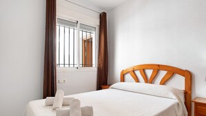 A neatly made bed with a wooden headboard, a window with bars, and a wooden nightstand to the right.
