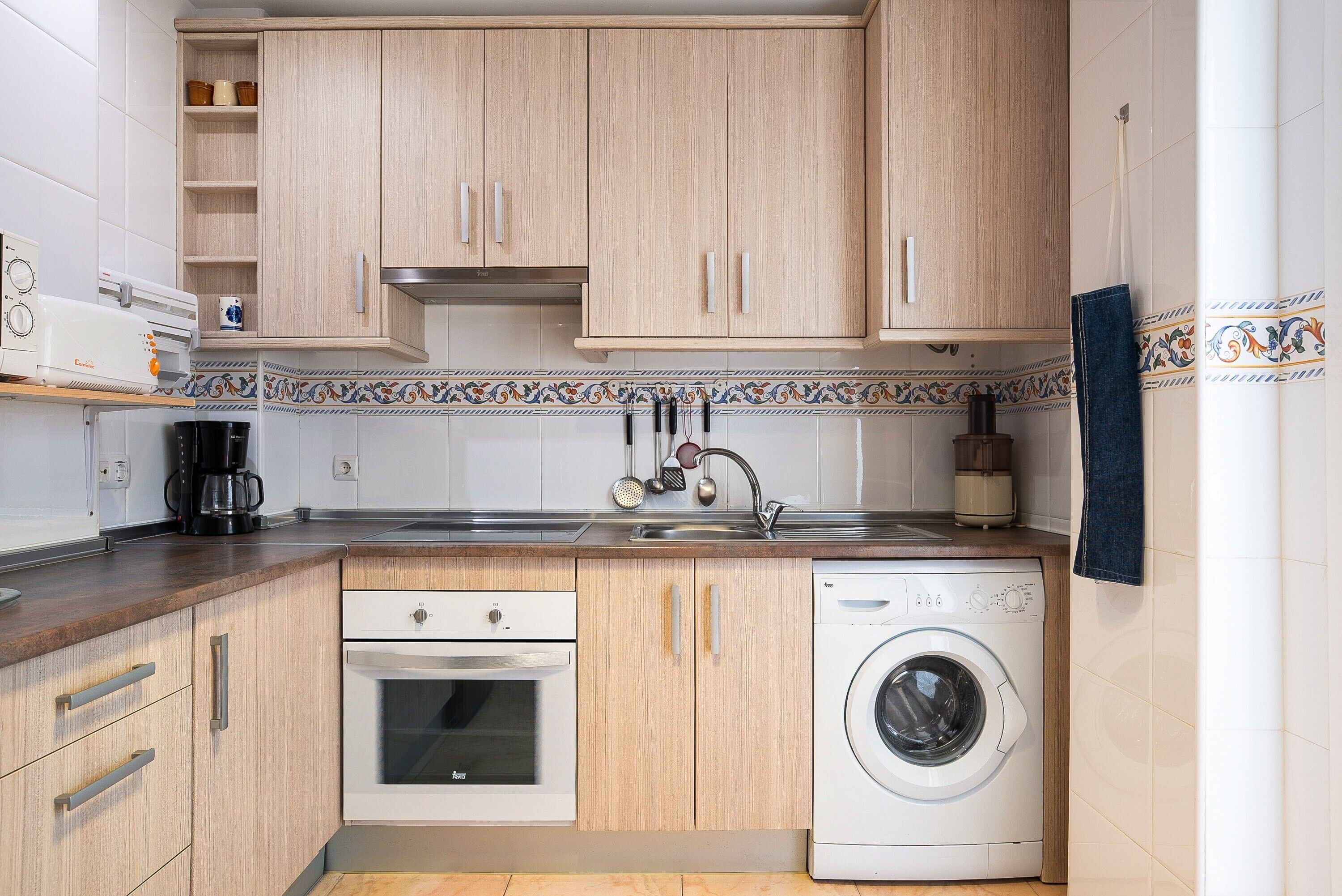 Een compacte keuken met houten kastjes, een ingebouwde oven, een wasmachine en een koffiezetapparaat.