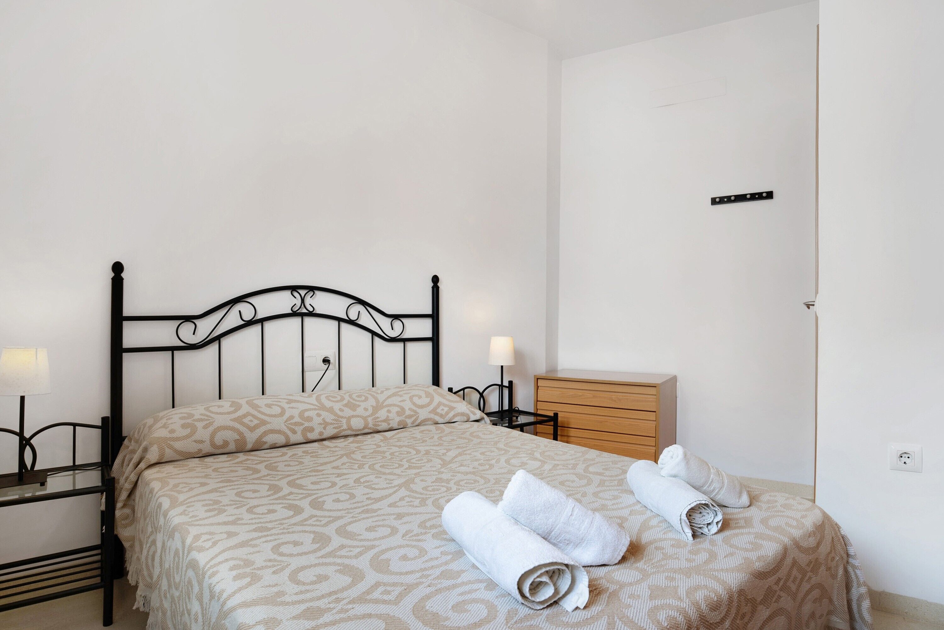 Ein ordentlich bezogenes Bett mit einer gemusterten Tagesdecke, ein Nachttisch mit Lampe, eine Holzkommode und ein an der Wand befestigter Lichtschalter.