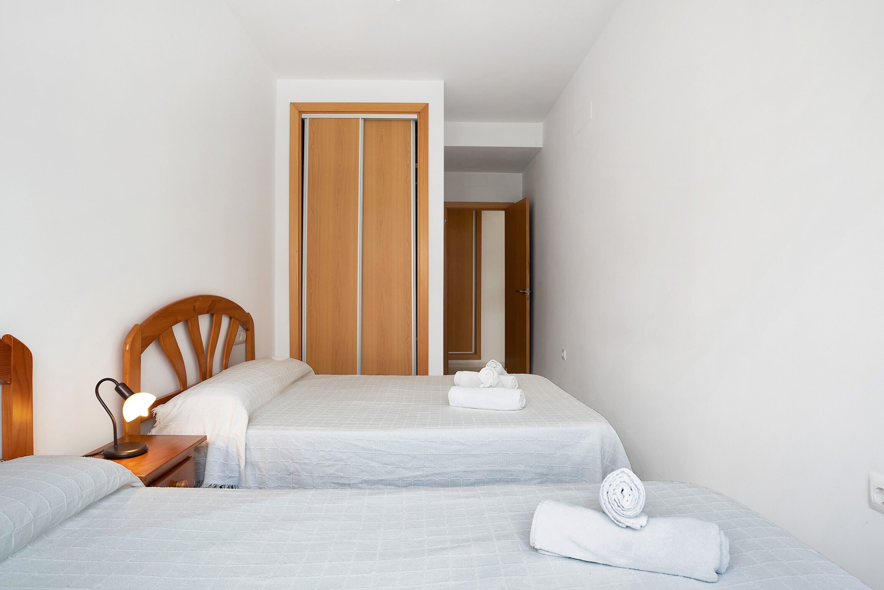 A hotel room with two beds, a wooden wardrobe, a nightstand with a lamp, and a bathroom door.