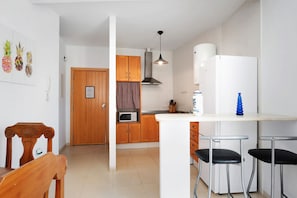 Een keuken met houten kastjes, een magnetron, een spoelbak en een witte koelkast. Er is een houten eettafel met stoelen en een hangende lamp.
